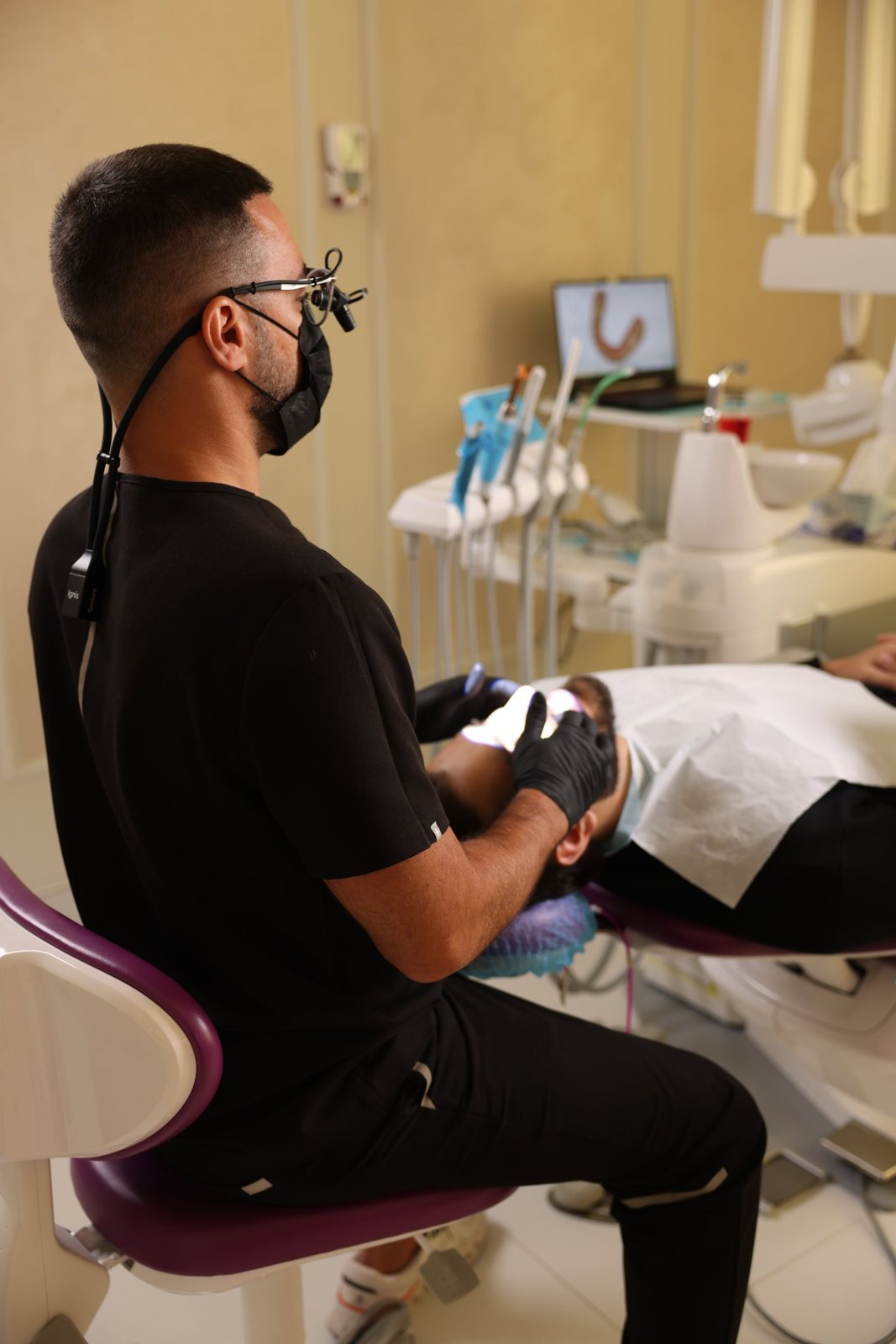 Dentist with loupes treating a patient, a digital scan of a dental arch on a laptop behind, Dubai